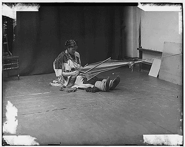 Black and white photograph of We-Wha, a Zuni two spirit, weaving while sitting on the ground