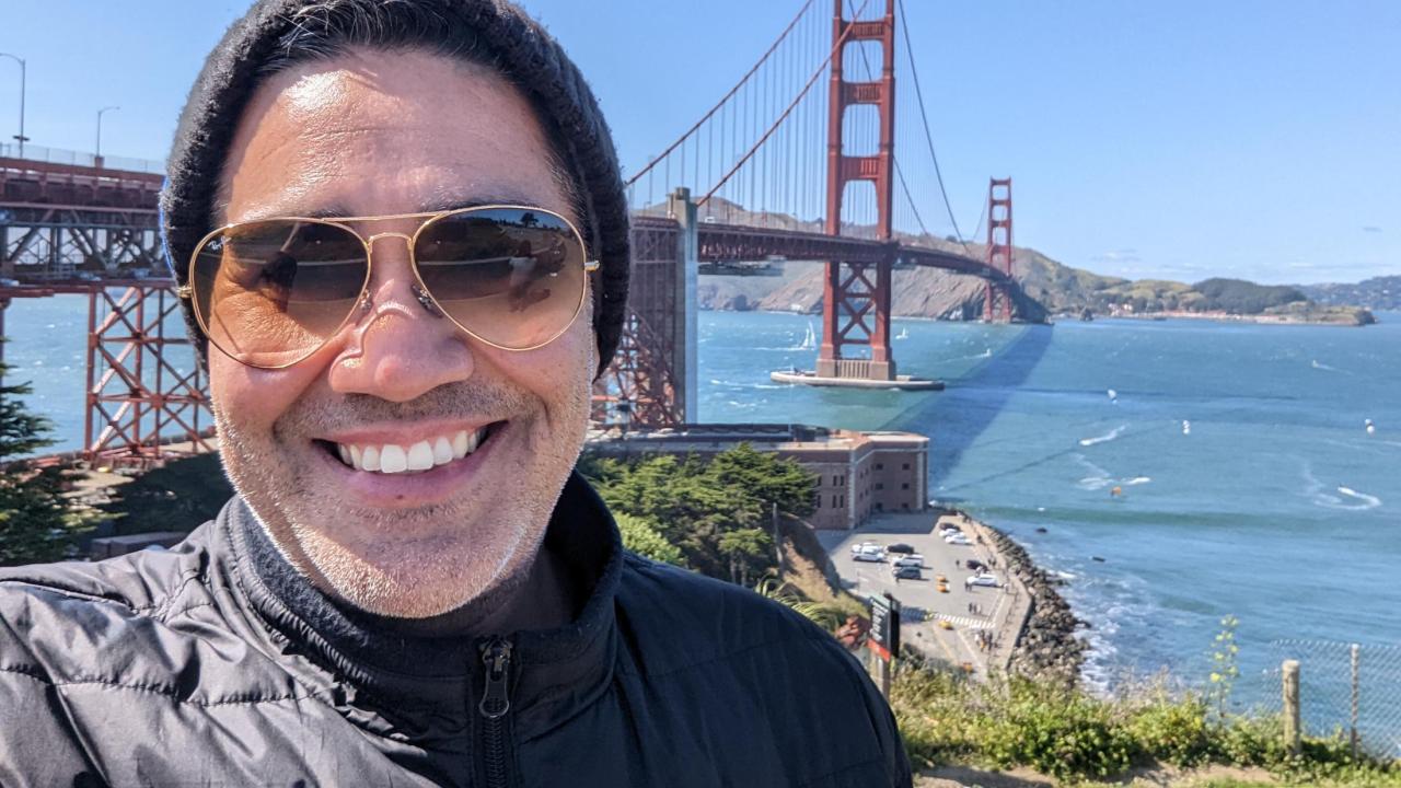 Jason Muniz stands in front of the Golden Gate Bridge wearing a hat and sunglasses. He is smiling and posing for a selfie.