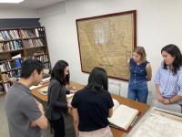 5 individuals lean over a table covered in neatly arranged historical documents paying particular attention to a ledger with yellowed pages. One individual, Professor Cecilia Tsu, compares her printed notes to the ledger.
