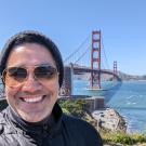 Jason Muniz stands in front of the Golden Gate Bridge wearing a hat and sunglasses. He is smiling and posing for a selfie.